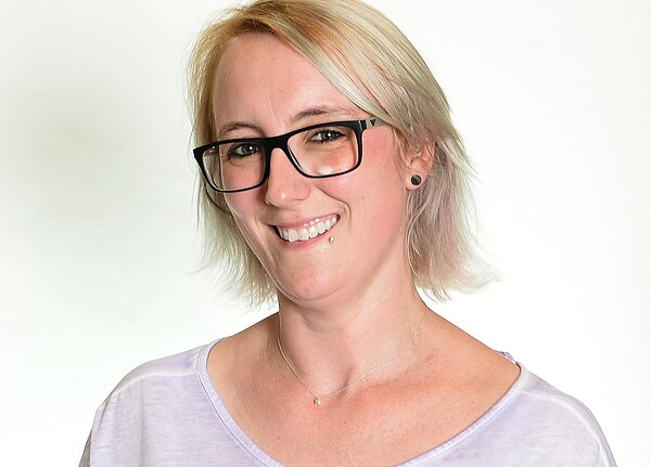 Porträt einer lächelnden Person mit kurzem blondem Haar, schwarzen rechteckigen Brillen, kleinem Lippenpiercing und Ohrstecker. Sie trägt ein helles T‑Shirt und eine feine Halskette. Neutraler, heller Hintergrund.