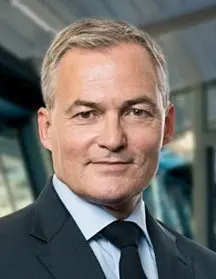 Ein Mann mittleren Alters mit grauen Haaren und einem freundlichen Ausdruck trägt einen schwarzen Anzug und eine hellblaue Krawatte. Er steht vor einem verschwommenen Hintergrund, der einen modernen Raum suggeriert.