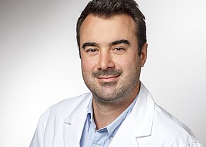 Dr. med. Milan Misovic Porträt eines Mannes mit kurzem dunklem Haar und Bartstoppeln, der freundlich in die Kamera blickt. Er trägt einen weißen Kittel vor neutralem Hintergrund.