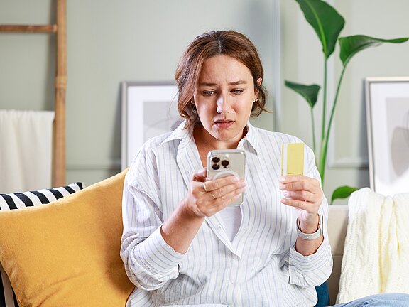 Eine besorgte Frau in einem gestreiften Hemd sitzt auf einem Sofa. Sie hält ein Telefon in der einen Hand und eine gelbe Karte in der anderen. Ihr Gesichtsausdruck zeigt Besorgnis, während sie auf den Bildschirm schaut.