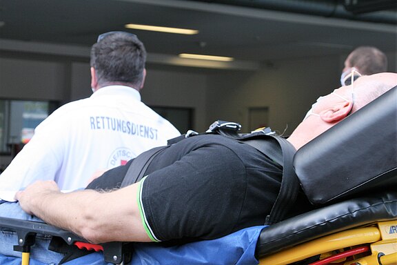 Ein Mann liegt auf einer Tragbahre, während ein Rettungsdienstmitarbeiter im Hintergrund steht. Der Mann trägt eine Maske und hat eine schwarze Oberbekleidung mit einem grünen Streifen. Die Szene wirkt hektisch, möglicherweise in einem Krankenhaus oder einer Rettungsstation.