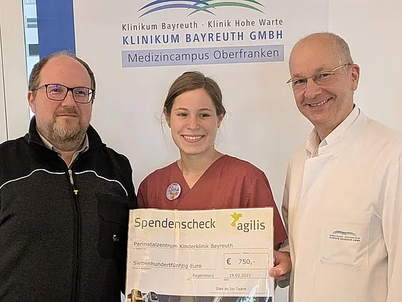 Drei Personen präsentieren einen Spendenscheck über 750 Euro. Die Spende von agilis geht an das Perinatalzentrum der Kinderklinik Bayreuth. Aufnahme im Klinikum Bayreuth; die mittlere Person in medizinischer Kleidung hält den Scheck, alle lächeln.