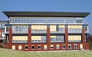 Ein modernes, dreigeschossiges Gebäude mit großen Fenstern und einem flachen Dach. Die Fassade ist rot mit gelben Jalousien und klaren Gebäudestrukturen. Der Himmel ist blau und sonnig, was dem Gebäude ein einladendes Aussehen verleiht.