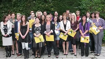 Eine Gruppe von etwa 20 Personen steht zusammen im Freien, lächelt und hält Urkunden in den Händen. Die Anwesenden sind frauen- und männergemischt, in verschiedenen Kleidungsstilen, und scheinen an einer Feier oder Zeremonie teilzunehmen. Im Hintergrund sind Pflanzen zu sehen.