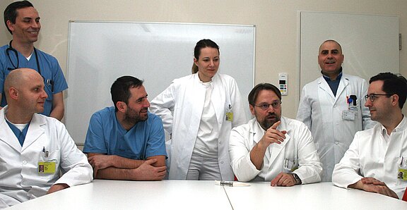 Mehrere Ärztinnen und Ärzte in weißen Kitteln und OP-Kleidung sitzen und stehen um einen Tisch. Sie führen eine Teambesprechung; eine Person spricht und gestikuliert, die anderen hören zu. Klinische Umgebung, Whiteboard im Hintergrund.