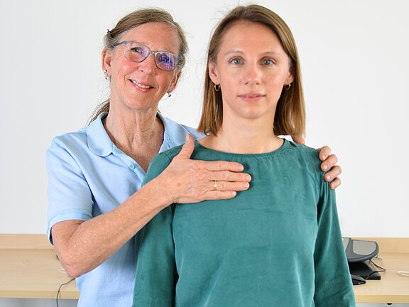 Eine ältere Frau steht hinter einer jüngeren und legt ihr eine Hand auf die Schulter, die andere auf den Brustbereich. Beide blicken ruhig in die Kamera. Die Geste wirkt unterstützend, als ob sie eine Untersuchung, Anleitung oder Therapie zur Körperwahrnehmung oder Atmung durchführt.