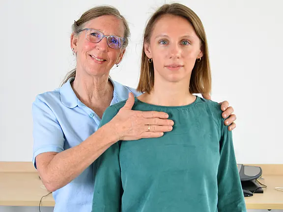 Eine ältere Frau steht hinter einer jüngeren und legt ihr eine Hand auf die Schulter, die andere auf den Brustbereich. Beide blicken ruhig in die Kamera. Die Geste wirkt unterstützend, als ob sie eine Untersuchung, Anleitung oder Therapie zur Körperwahrnehmung oder Atmung durchführt.
