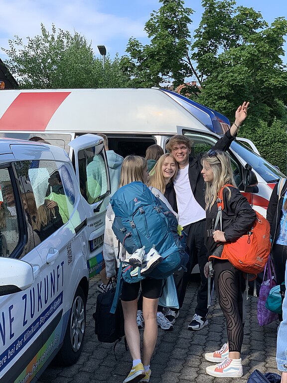 Gruppe junger Menschen mit Rucksäcken steigt gut gelaunt in einen Kleinbus. Ein junger Mann winkt und lächelt. Die Szene wirkt wie die Abfahrt zu einer gemeinsamen Reise oder Freizeit.