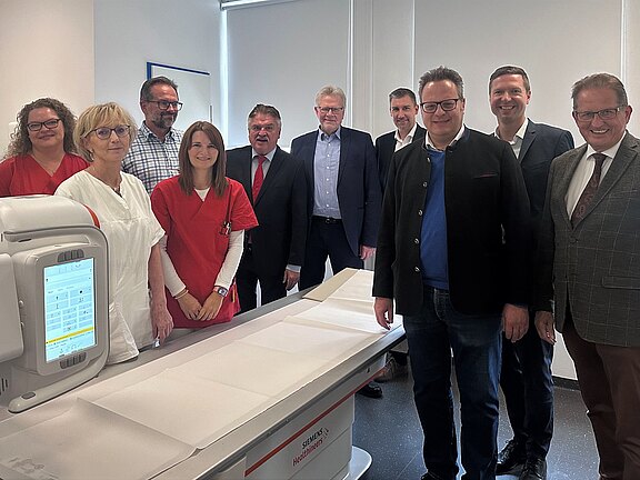 Gruppenfoto von Mitarbeitern und Vertretern in einem modernen medizinischen Raum. Sie stehen um ein Gerät der Marke Siemens Healthineers, das auf einem Behandlungstisch platziert ist. Die Personen sind freundlich und tragen sowohl Arbeitskleidung als auch formelle Kleidung.