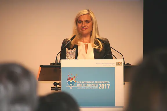 Eine blonde Frau spricht am Rednerpult bei einer Veranstaltung mit der Aufschrift „Der Bayerische Gesundheits- und Pflegepreis 2017“. Mikrofone vor ihr, Glas Wasser auf dem Pult. Publikum im Vordergrund unscharf.