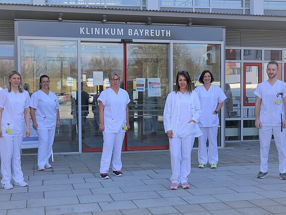 Sechs Mitarbeitende in weißer Krankenhauskleidung stehen vor dem Haupteingang des Klinikum Bayreuth und blicken in die Kamera.
