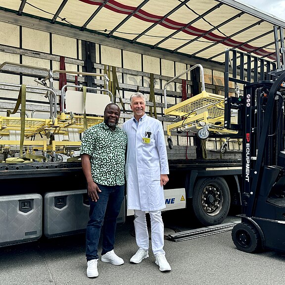 Zwei Männer stehen vor einem Lieferwagen, der mit medizinischer Ausrüstung beladen ist. Der Mann links trägt ein grünes Hemd mit Muster, der rechts einen weißen Arztkittel. Im Hintergrund sind Krankenhausbetten sichtbar, die zur Entladung bereitstehen.