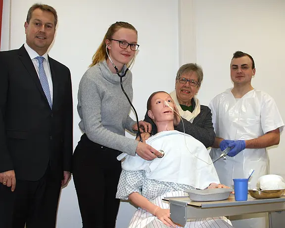 In einem Schulungsraum übt eine Pflegeschülerin mit Stethoskop an einer realistischen Patientensimulationspuppe. Neben ihr stehen eine Ausbilderin, ein weiterer Pflegeschüler in Schutzkleidung und eine Person im Anzug. Das Bild vermittelt Pflegeausbildung und praktische Übung am Simulator.