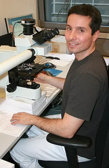Ein Mann sitzt an einem Tisch, der mit einem Mikroskop und wissenschaftlichen Unterlagen ausgestattet ist. Er lächelt freundlich in die Kamera und trägt ein dunkelgraues T-Shirt. Im Hintergrund sind Fenster zu sehen, durch die Tageslicht fällt.