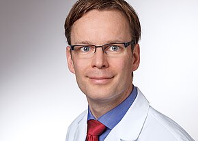 Dr. Richard Sommerer Porträt einer lächelnden Person mit Brille in weißem Kittel, blauem Hemd und roter Krawatte vor neutralem Hintergrund. Kopf-und-Schultern-Aufnahme, direkter Blick in die Kamera.