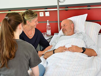 Eine Frau spricht freundlich mit einem Mann, der im Bett liegt. Eine junge Frau sitzt daneben und hört zu. Der Mann hat einen nachdenklichen Gesichtsausdruck, während die Frauen lächeln. Die Atmosphäre wirkt warm und unterstützend, in einem modernen Krankenhauszimmer.