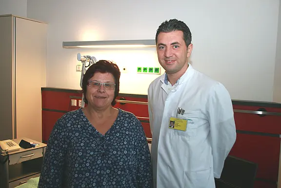 Eine Frau und ein Arzt im weißen Kittel stehen in einem Untersuchungsraum und lächeln; klinische Umgebung mit Geräten im Hintergrund.