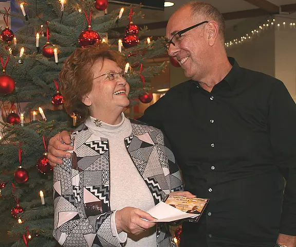 Zwei Erwachsene stehen vor einem geschmückten Weihnachtsbaum. Sie schauen sich lächelnd an, der Mann hat den Arm um die Frau gelegt. Die Frau hält einen Umschlag und eine Karte in der Hand, als ob sie ein Geschenk oder Glückwünsche erhält.