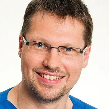 Ein lächelnder Mann mit Brille trägt ein blaues Oberteil. Sein Gesicht zeigt ein freundliches und offenes Ausdruck. Der Hintergrund ist neutral und hell gestaltet.