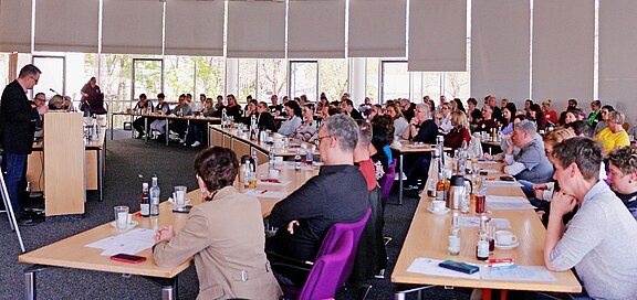 Großer Tagungsraum: Viele Menschen sitzen an U-förmigen Tischen und hören einem Redner am Pult zu; fokussierte Atmosphäre einer Präsentation oder Versammlung.