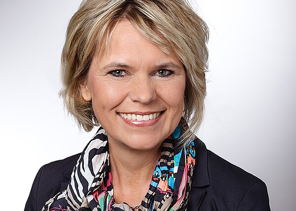 Porträtfoto einer lächelnden Frau mit kurzem blondem Haar. Sie trägt einen dunklen Blazer und einen bunten, gemusterten Schal. Heller, neutraler Hintergrund; wirkt wie ein professionelles Geschäfts- oder Bewerbungsfoto.