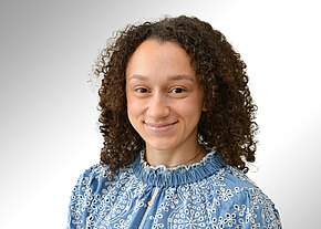 Bria-Estella Johnson Porträt einer lächelnden Person mit dunkelbraunen Locken, leicht nach rechts gewandt. Sie trägt eine hellblaue, reich bestickte Bluse mit hohem Kragen. Neutraler, heller Hintergrund.