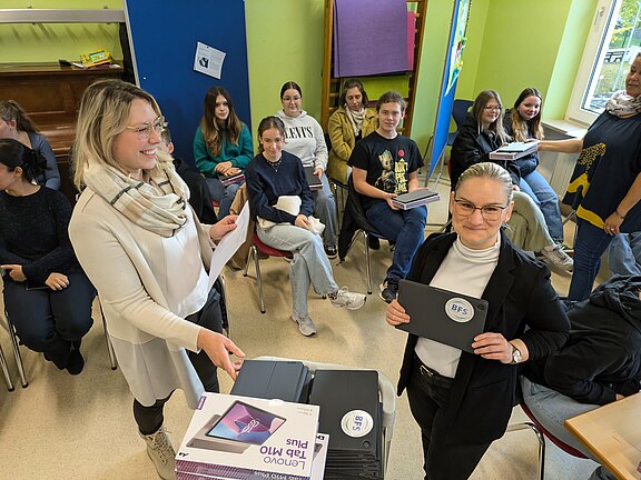 In einem Klassenraum verteilen zwei Lehrerinnen Tablets an eine Gruppe sitzender Schüler. Die Lernenden halten Geräte oder Verpackungen in den Händen und lächeln. Im Vordergrund zeigt eine Frau ein Tablet mit Schullogo. Die Szene wirkt wie eine offizielle Übergabe neuer Unterrichtstablets.