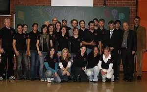 Eine Gruppe von etwa 20 Personen steht in einem Raum, alle tragen schwarze T-Shirts. Im Hintergrund sind bunte Ballons und ein beschriftetes Stück Kreidetafel zu sehen. Einige Personen sind in formeller Kleidung. Die Stimmung wirkt freundlich und gemeinschaftlich.