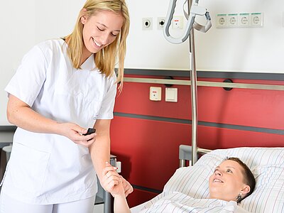 Pflegekraft im weißen Kittel lächelt und misst mit einem Fingerclip die Sauerstoffsättigung eines lächelnden Patienten, der im Krankenhausbett liegt. Die Pflegekraft hält die Hand.