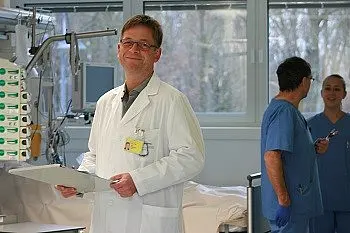 Arzt im weißen Kittel steht in einem Klinikraum, lächelt und hält ein Klemmbrett. Im Hintergrund sprechen zwei Pflegekräfte in blauen Kitteln neben medizinischen Geräten. Helle Fenster im Raum.