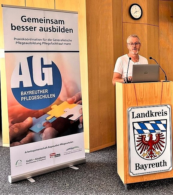 Eine Person spricht am Rednerpult des Landkreises Bayreuth. Daneben ein Roll‑Up mit dem Logo „AG Bayreuther Pflegeschulen“ und dem Slogan „Gemeinsam besser ausbilden“ zur Praxiskoordination der generalistischen Pflegeausbildung. Fokus auf Vorstellung einer Bildungsinitiative im Pflegebereich.