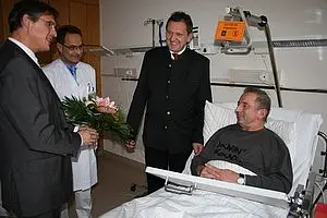 Vier Männer stehen im Krankenhauszimmer. Ein Mann im Bett hält einen Strauß Blumen. Die anderen drei, einer in Arztkleidung, sprechen freundlich miteinander. Der Mann im Bett hat ein entspanntes Lächeln und scheint sich über den Besuch zu freuen.