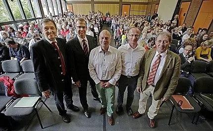 Eine Gruppe von fünf Männern steht im Vordergrund, während im Hintergrund eine große Menschenmenge in einem Raum sitzt. Die Atmosphäre wirkt informell und lebhaft, was auf eine Veranstaltung oder eine Ansprache hindeutet.
