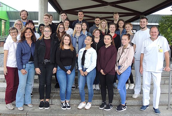 Eine Gruppe von etwa 25 Personen steht auf einer Treppe vor einem Gebäude. Sie lächeln in die Kamera, einige tragen Freizeitkleidung, andere Berufsbekleidung. Die Szene vermittelt einen freundlichen und gemeinschaftlichen Eindruck.