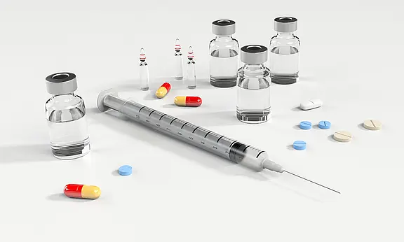 Eine liegende Spritze mit aufgesetzter Nadel, umgeben von mehreren durchsichtigen Medikamentenfläschchen sowie verstreuten Tabletten und Kapseln in verschiedenen Farben. Symbolbild für Medizin, Impfung oder Behandlung.