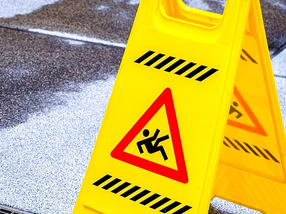 Ein gelbes Warnschild steht auf einem nassen Boden. Es zeigt ein Symbol für Rutschgefahr und warnt Passanten, vorsichtig zu sein. Der Boden ist uneben und weist eine klare Feuchtigkeit auf, was das Risiko von Stürzen erhöht.