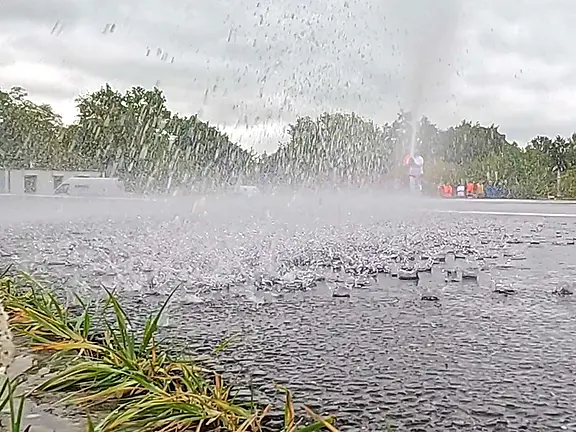 Ein Wasserstrahl spritzt aus dem Boden, während sich der Regen auf der asphaltierenden Fläche verteilt. Im Hintergrund sind grüne Bäume und verschwommene Figuren zu erkennen, die sich in einer leicht nebligen Umgebung bewegen. Der Himmel ist bewölkt.