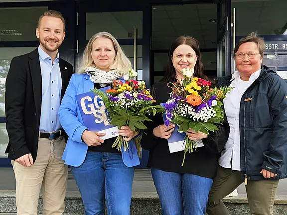 Vier Personen stehen auf einer Treppe vor einem Gebäude. Zwei Frauen halten bunte Blumensträuße, während ein Mann und eine Frau lächeln. Alle wirken erfreut und stehen in einer geselligen Atmosphäre.