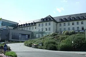 Ein mehrstöckiges Gebäude mit moderner Glasarchitektur ist im Vordergrund zu sehen. Im Hintergrund ist ein traditionellerer Gebäudeteil mit einem Satteldach. Um das Gebäude herum gibt es eine grüne Landschaft mit Blumen und Büschen.