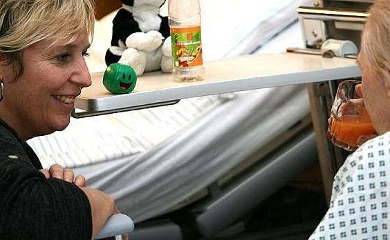 Zwei Frauen sitzen sich in einem Krankenhauszimmer gegenüber. Die eine Frau lächelt und schaut aufmerksam, während die andere einen Becher hält. Zwischen ihnen steht ein Tisch mit einer Flasche und einem Spielzeug. Im Hintergrund ist ein Bett zu sehen.