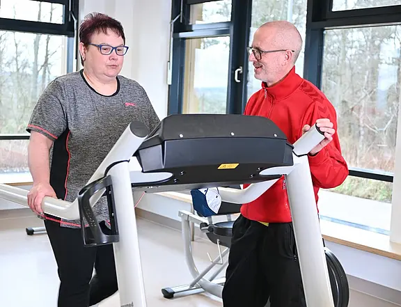 In einem hellen Fitnessraum trainiert eine Frau auf einem Laufband. Ein Trainer steht neben ihr, spricht mit ihr und kontrolliert die Einstellungen. Die Szene zeigt betreutes Ausdauertraining.