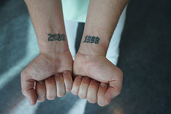 Zwei geschlossene Fäuste zeigen die Innenseiten der Handgelenke. Auf dem linken Handgelenk steht tätowiert „2019“, auf dem rechten „1998“, in schwarzer, gotischer Schrift. Fokus liegt auf den Jahreszahlen.