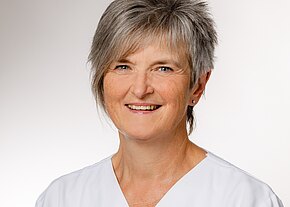 Christine Herold Eine lächelnde Frau mit kurzen, grauen Haaren trägt eine weiße Berufsbekleidung. Sie hat einen freundlichen Gesichtsausdruck und schaut direkt in die Kamera. Der Hintergrund ist einfarbig und erzeugt einen professionellen Eindruck.
