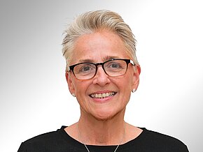 Alexandra Goldbaum-Carter Porträt einer lächelnden Frau mit kurzen, grauen Haaren und Brille. Sie trägt ein schwarzes Oberteil und Ohrstecker. Der Hintergrund ist hell und weich verschwommen.