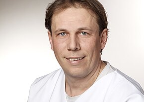 Station 6a Porträt eines Mannes in weißer Arbeitskleidung vor neutralem Hintergrund, der direkt in die Kamera blickt und leicht lächelt.