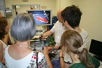 Eine Gruppe von Menschen steht um einen Arzt, der ihnen ein medizinisches Bild auf einem Monitor zeigt. Der Arzt erklärt etwas, während die anderen interessiert zuhören und einige Haltegeräte in den Händen halten.