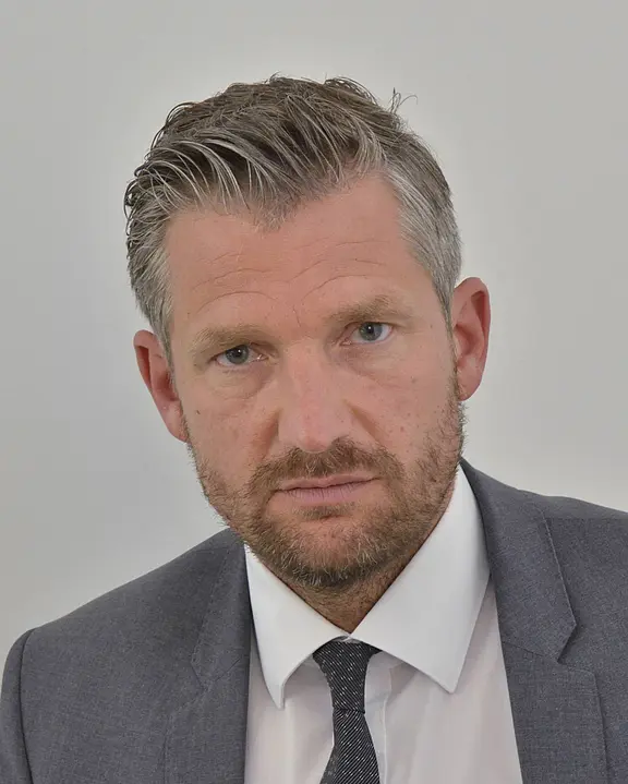 Porträt eines Mannes in grauem Anzug mit weißem Hemd und dunkler Krawatte. Er blickt ernst direkt in die Kamera; kurzes, graumeliertes Haar und Bartstoppeln. Neutraler, heller Hintergrund.