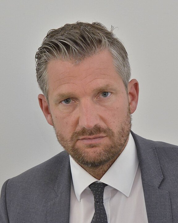 Porträt eines Mannes in grauem Anzug mit weißem Hemd und dunkler Krawatte. Er blickt ernst direkt in die Kamera; kurzes, graumeliertes Haar und Bartstoppeln. Neutraler, heller Hintergrund.