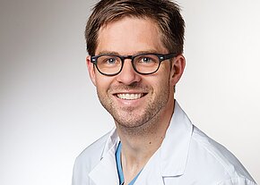 Porträt eines lächelnden Mannes mit Brille in weißem Arztkittel vor neutralem Hintergrund, Blick in die Kamera.