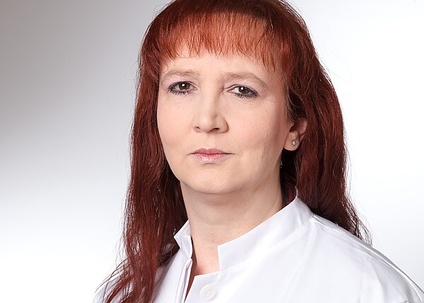Nahaufnahme einer erwachsenen Person mit rötlichen, schulterlangen Haaren und neutralem Blick. Sie trägt einen weißen Kittel vor hellem, neutralem Hintergrund.